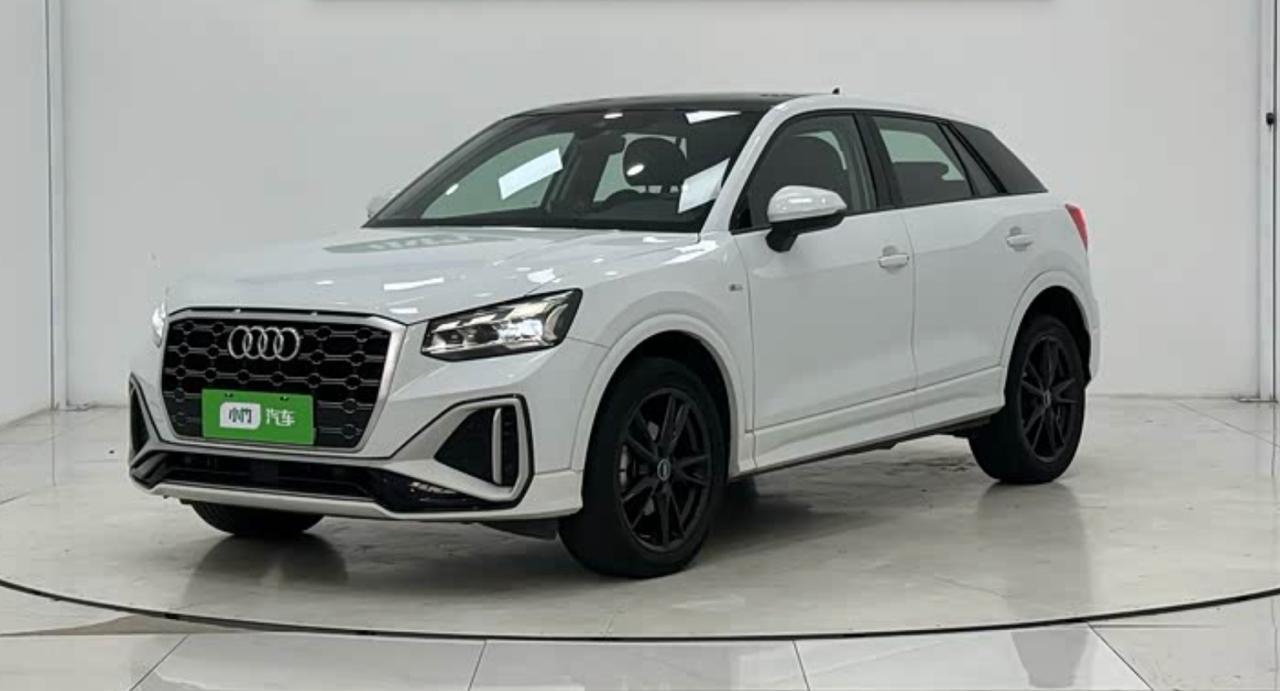 Белый Audi Q2L 2022 года с чёрной крышей и чёрными дисками на поворотном круге в салоне китайского дилера, вид спереди сбоку